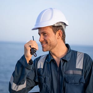 Cheerful young workman in protective uniform and hardhat speaking on walkie talkie radio set while working at sea port