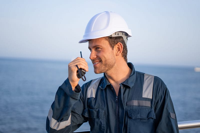 Cheerful young workman in protective uniform and hardhat speaking on walkie talkie radio set while working at sea port